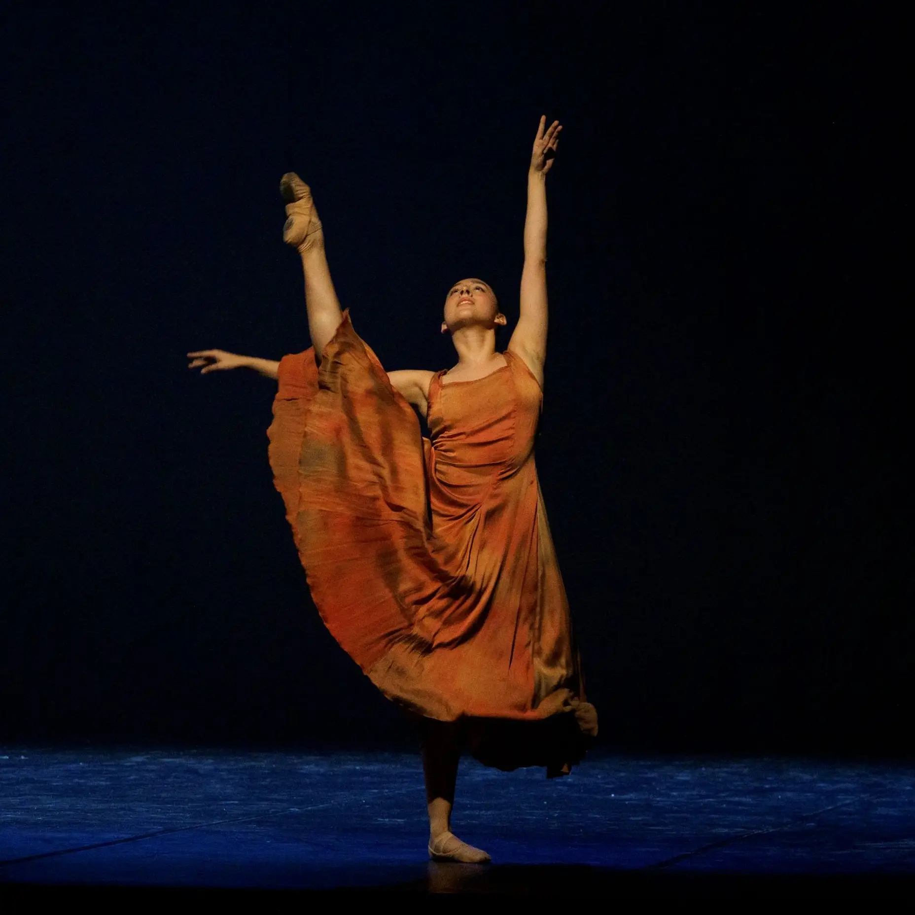 Une ballerine se tient sur scène, vêtue d'une robe orange. Elle tend une jambe vers le haut et lève les bras.
