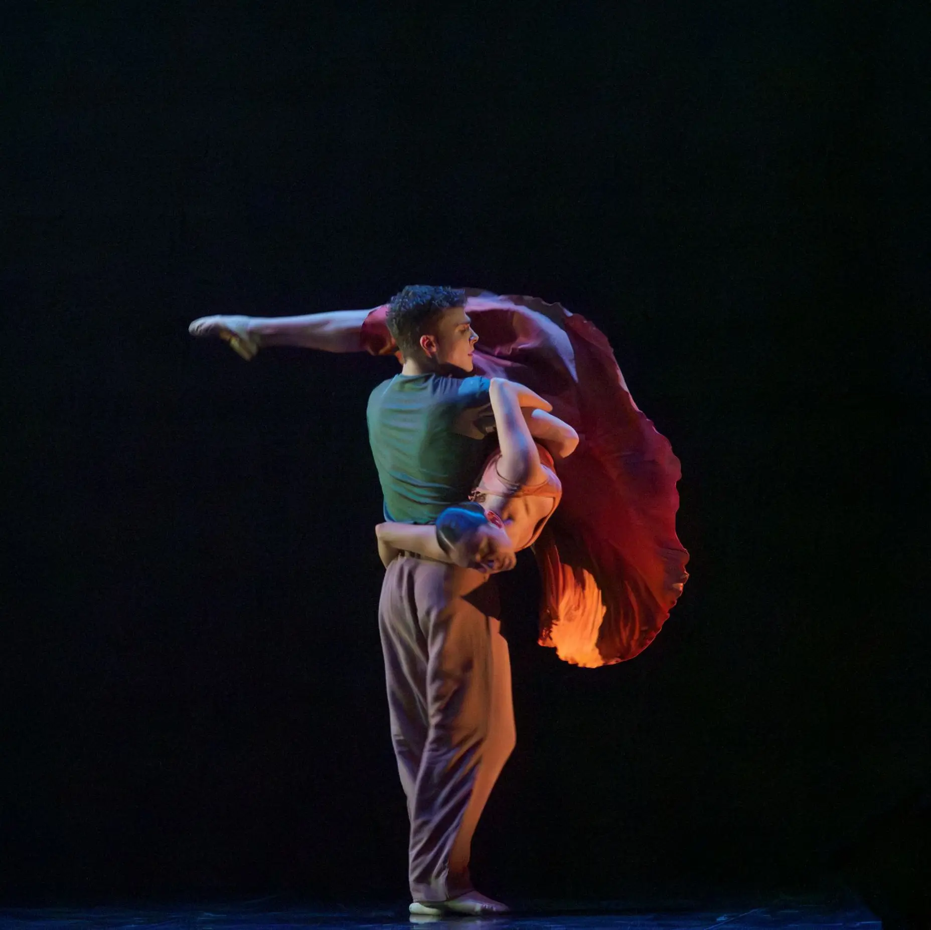 Une danseuse de ballet vêtue d'une robe rouge est soulevée par un danseur de ballet portant un t-shirt vert et un pantalon rouge. Elle passe entre ses bras et tend sa jambe par-dessus son épaule.