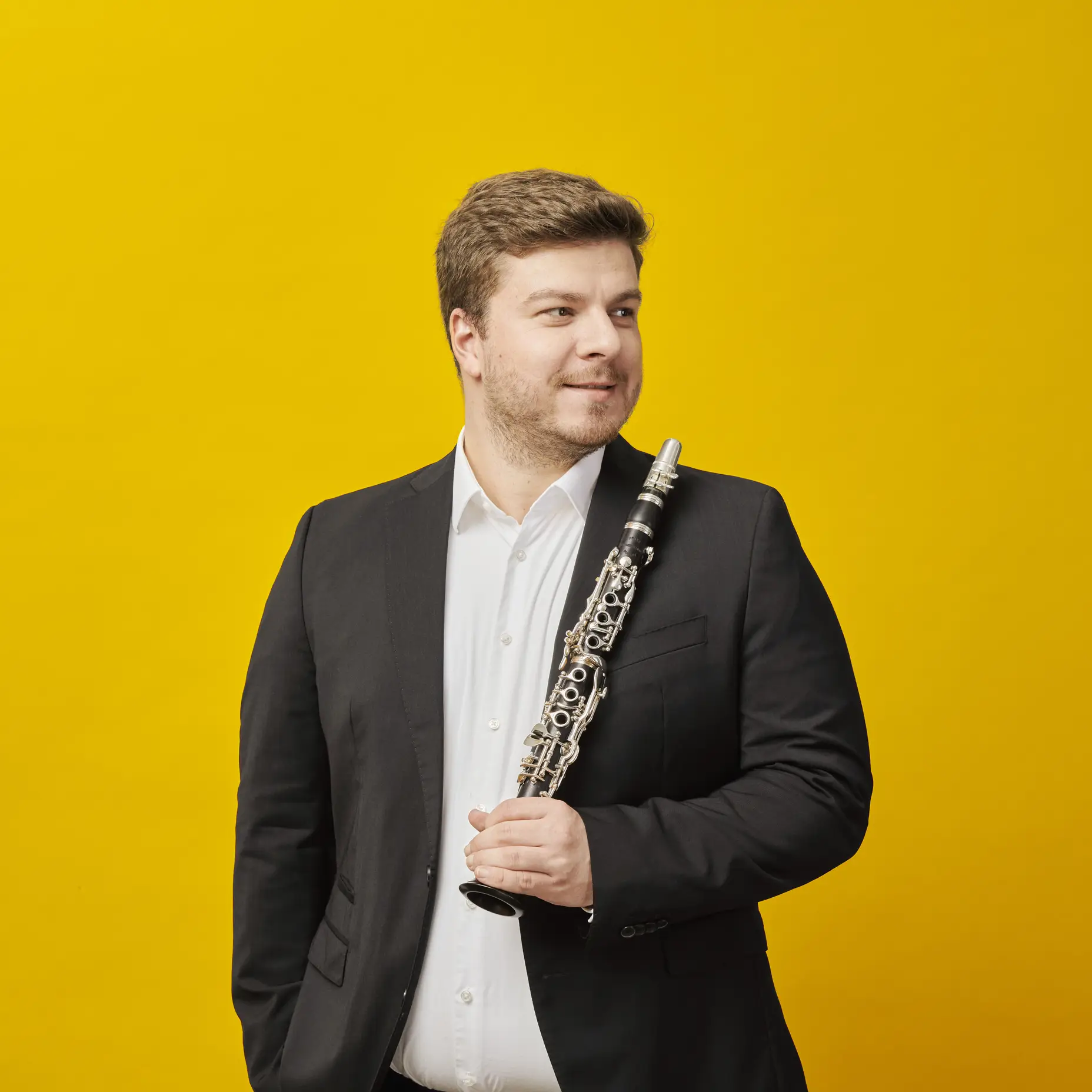 Ein Mann in einem schwarzen Anzug hält eine Oboe vor einem hellgelben Hintergrund. Er lächelt leicht.
