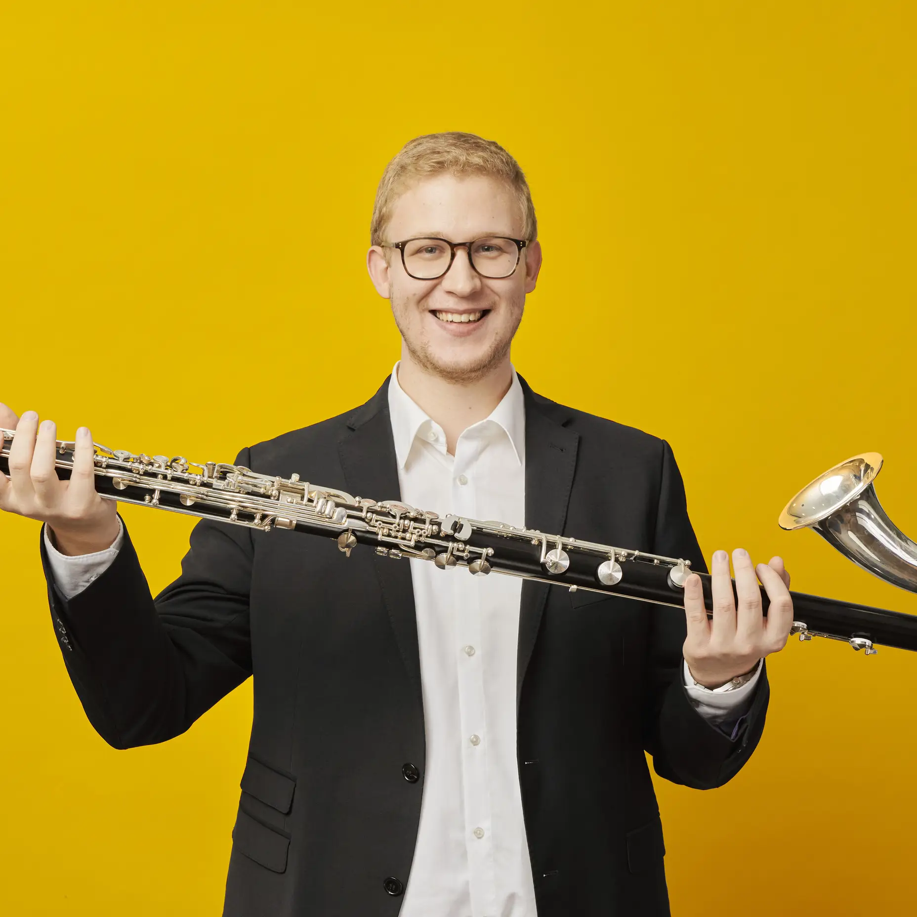 Ein Mann im Anzug hält ein großes Musikinstrument, ein Kontrabassklarinette, vor einem leuchtend gelben Hintergrund.