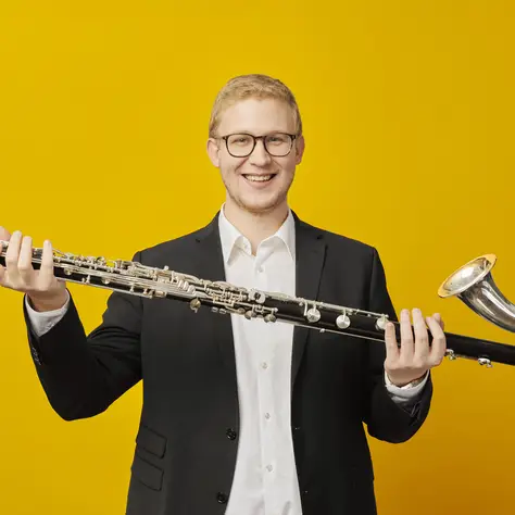 Ein Mann im Anzug hält ein großes Musikinstrument, ein Kontrabassklarinette, vor einem leuchtend gelben Hintergrund.
