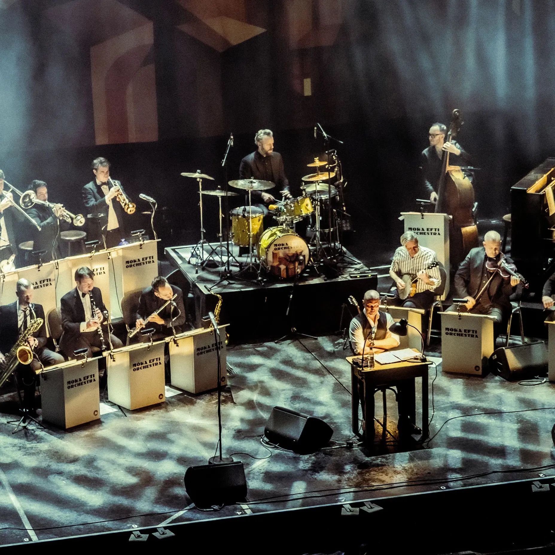 Eine Jazzband tritt auf einer Bühne auf. Mehrere Musiker spielen verschiedene Instrumente, darunter Saxophone, Trompeten und ein Schlagzeug.