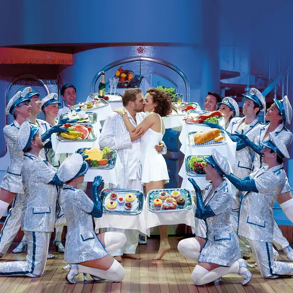 Szenenbild aus einem Musical, in dem ein Paar in der Mitte steht, umgeben von Tänzern in blauen Uniformen.
