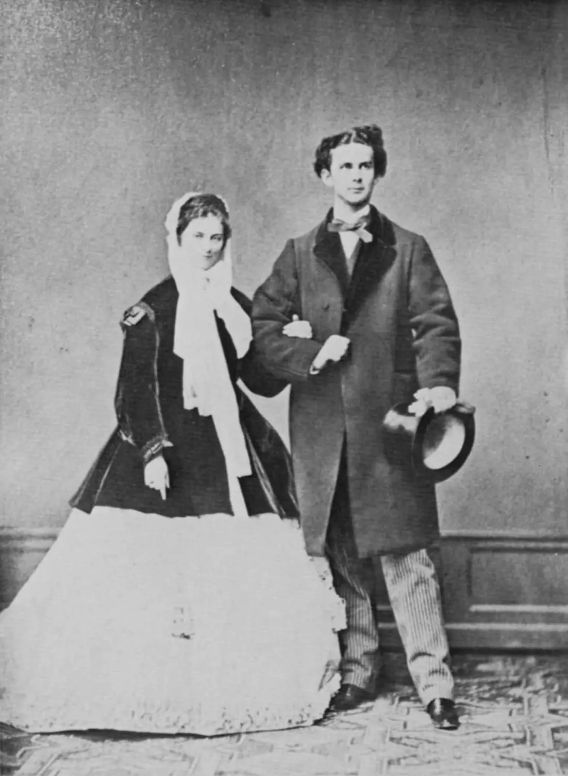 Un couple du 19ème siècle pose pour une photo. Ils portent des vêtements traditionnels victoriens. L'homme tient un chapeau.
