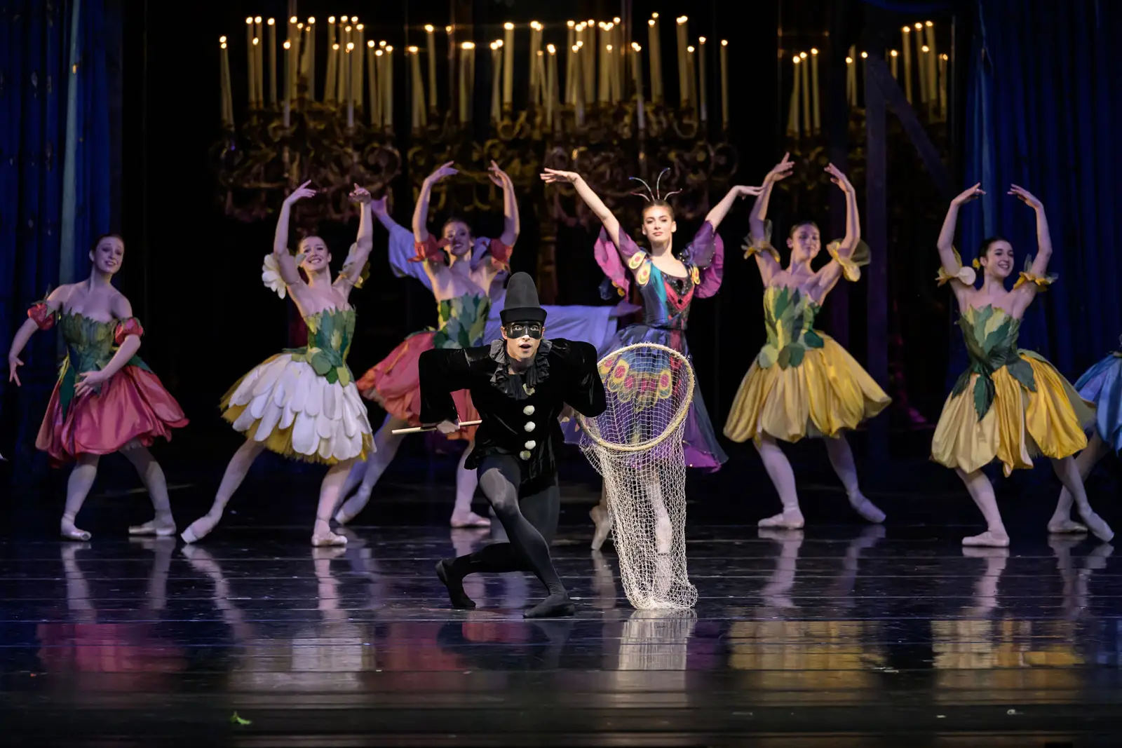 Danseurs de ballet en costumes colorés exécutent une chorégraphie élégante. Un danseur s'agenouille tandis que d'autres posent, avec un lustre en arrière-plan.