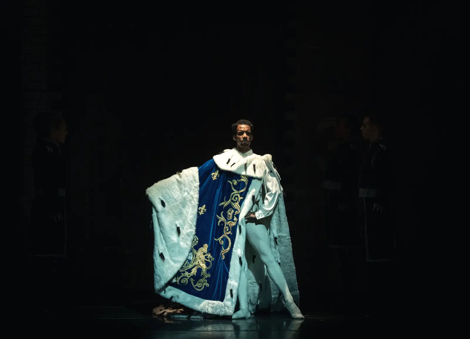 Un artiste en costume élaboré avec des broderies bleues et dorées sur scène dans une ambiance sombre.