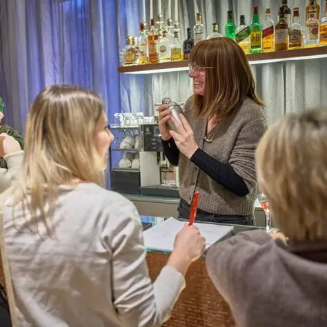 Eine Frau an einer Bar zeigt drei Frauen etwas. Es scheint eine interaktive und gesellige Umgebung zu sein.