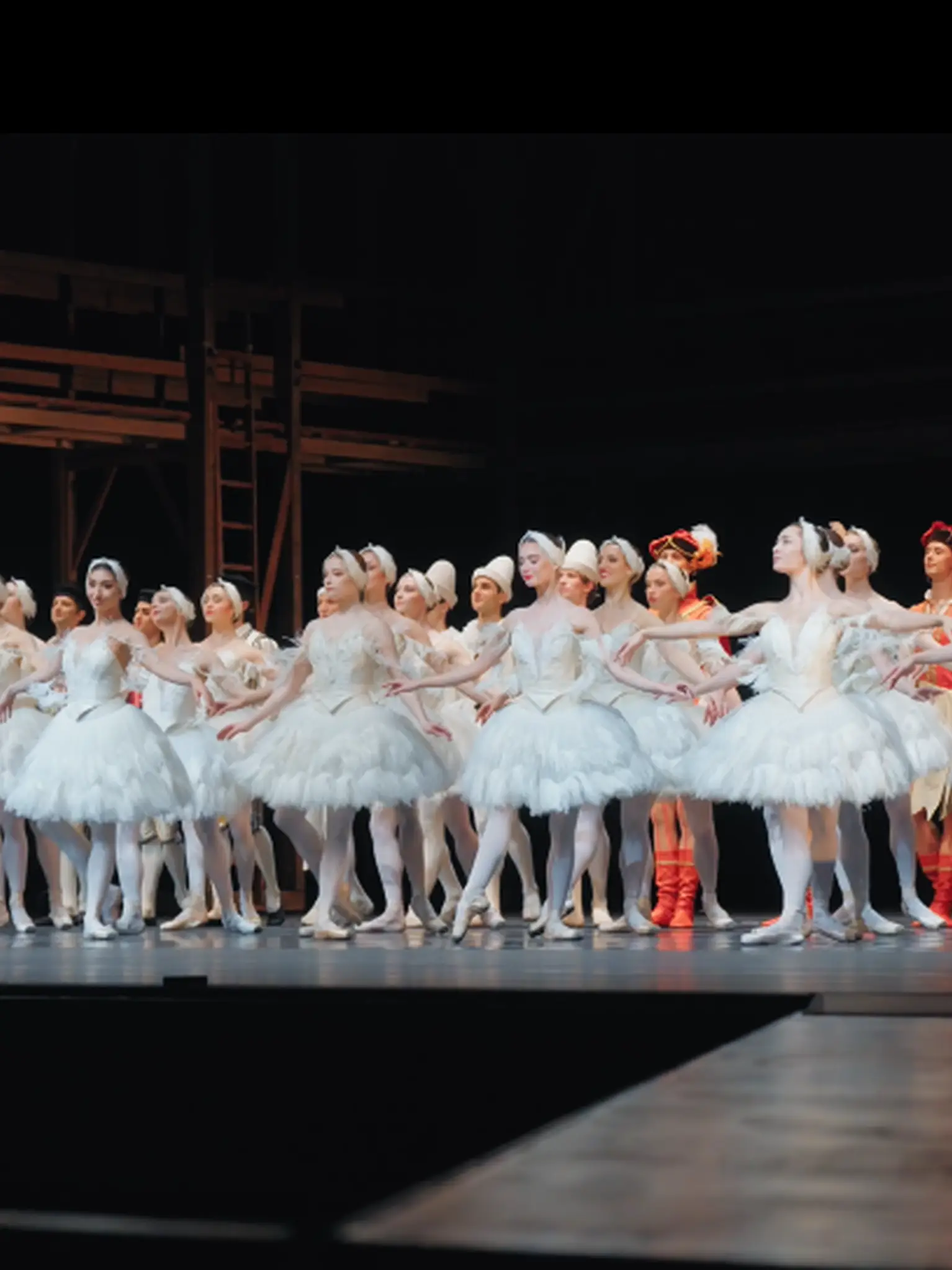 Eine Gruppe von Balletttänzerinnen in weißen Tutus steht auf einer Bühne, bereit für eine Aufführung. Der Hintergrund ist schlicht.