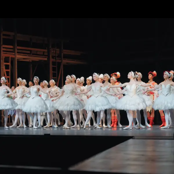 Eine Gruppe von Balletttänzerinnen in weißen Tutus steht auf einer Bühne, bereit für eine Aufführung. Der Hintergrund ist schlicht.