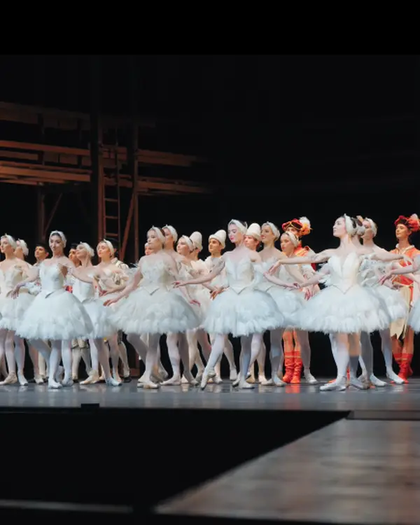 Eine Gruppe von Balletttänzerinnen in weißen Tutus steht auf einer Bühne, bereit für eine Aufführung. Der Hintergrund ist schlicht.