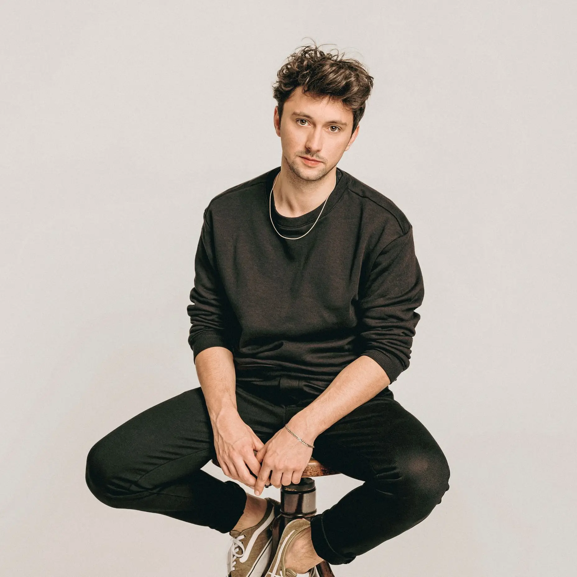 A man in casual black clothing sits on a stool against a neutral background, looking directly at the camera.