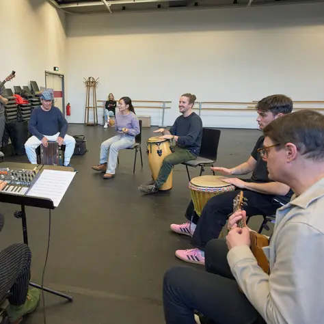 A group rehearsing in a room, with some playing instruments like guitar and drums, while another person conducts.