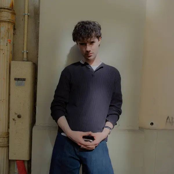 Eine Person mit lockigem Haar steht lässig an einer Wand, trägt ein dunkles Oberteil und blaue Jeans.