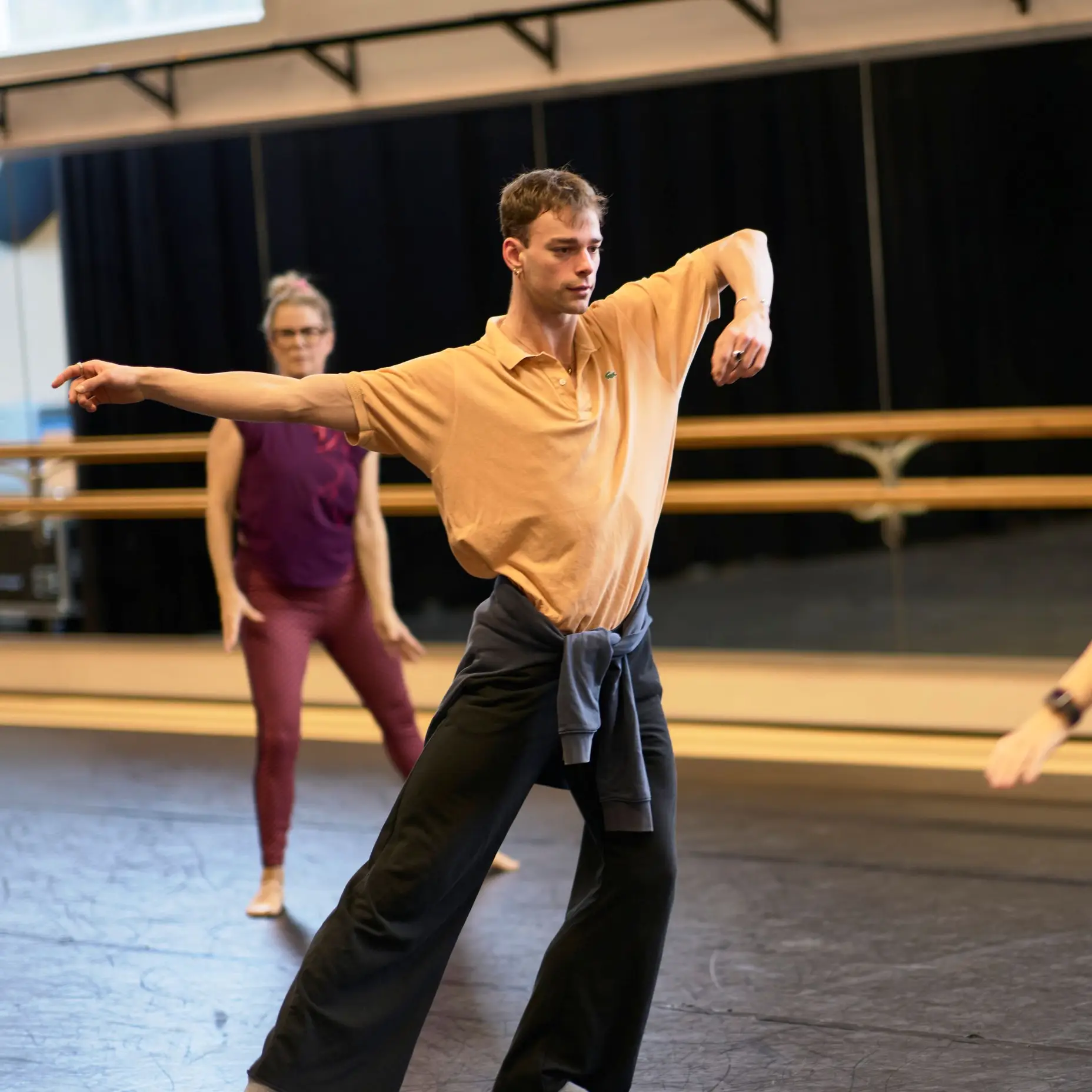 Drei Personen in einem Tanzstudio üben Bewegungen vor einem Spiegel. Ein Mann und eine Frau stehen einander gegenüber.