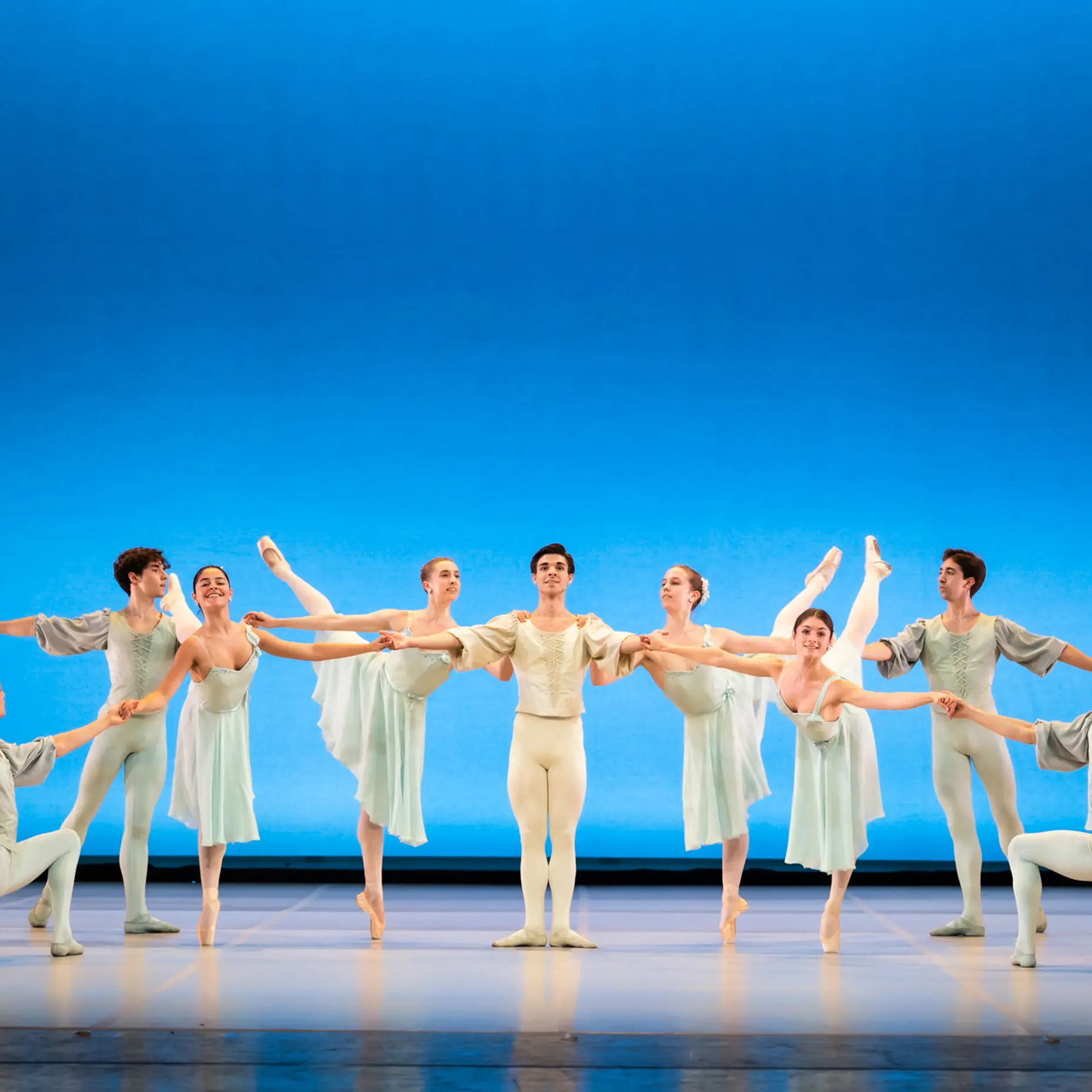 Acht Balletttänzer in Pastellkostümen führen eine klassische Choreographie auf einer Bühne mit blauem Hintergrund auf.