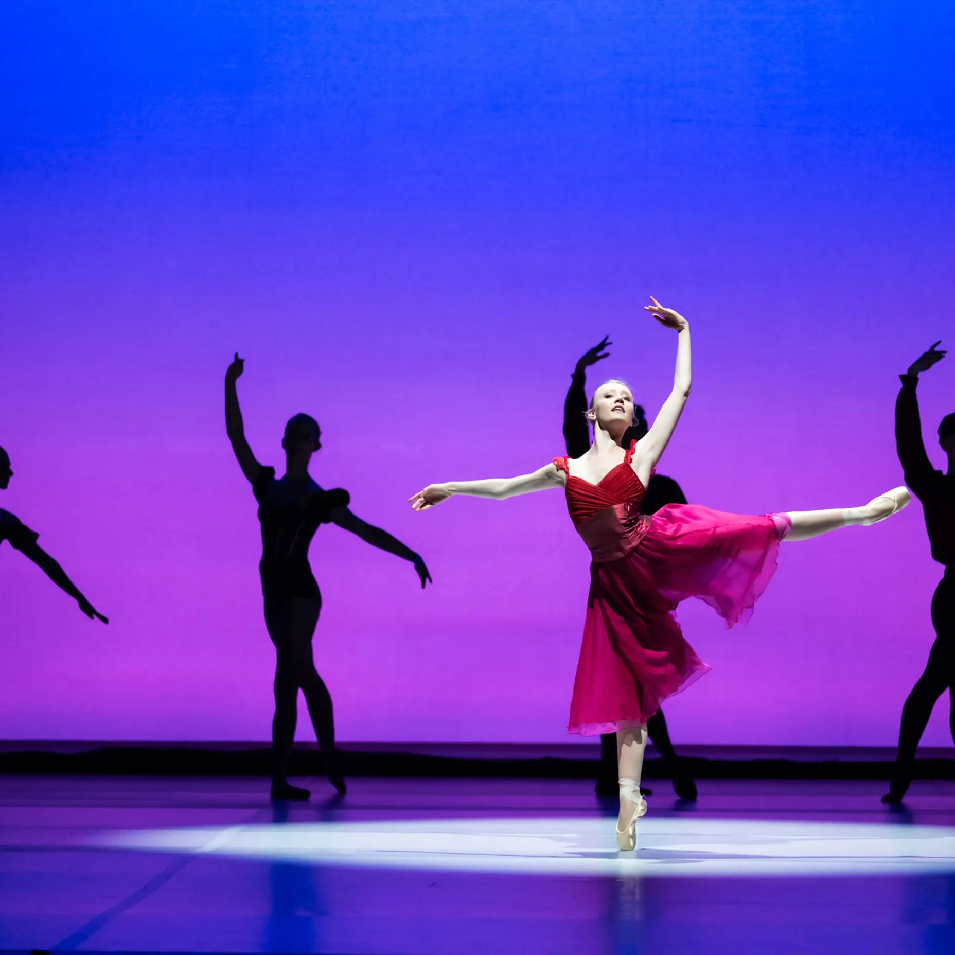 Balletttänzerin in rotem Kleid tanzt auf einer hell erleuchteten Bühne vor einem rosafarbenen Hintergrund, umgeben von vier dunkleren Tänzern.