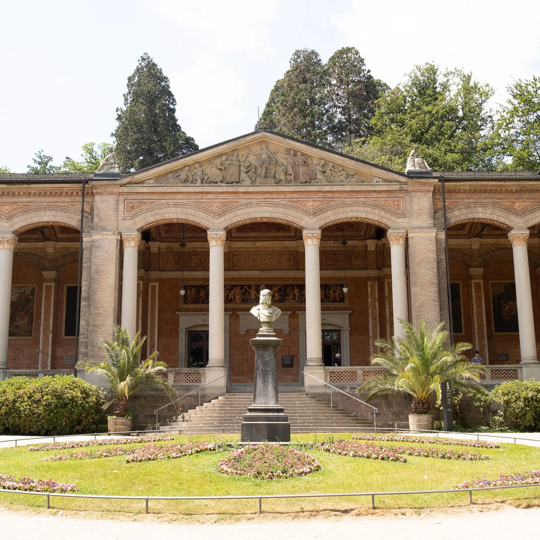 Un bâtiment avec une façade à colonnes et un buste central dans le jardin. Entouré d'arbres et de pelouses soignées.