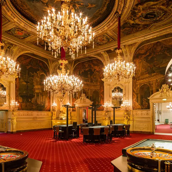 An opulent room with red carpets and grand chandeliers hanging from the ceiling. The walls are richly decorated. Luxurious setting.