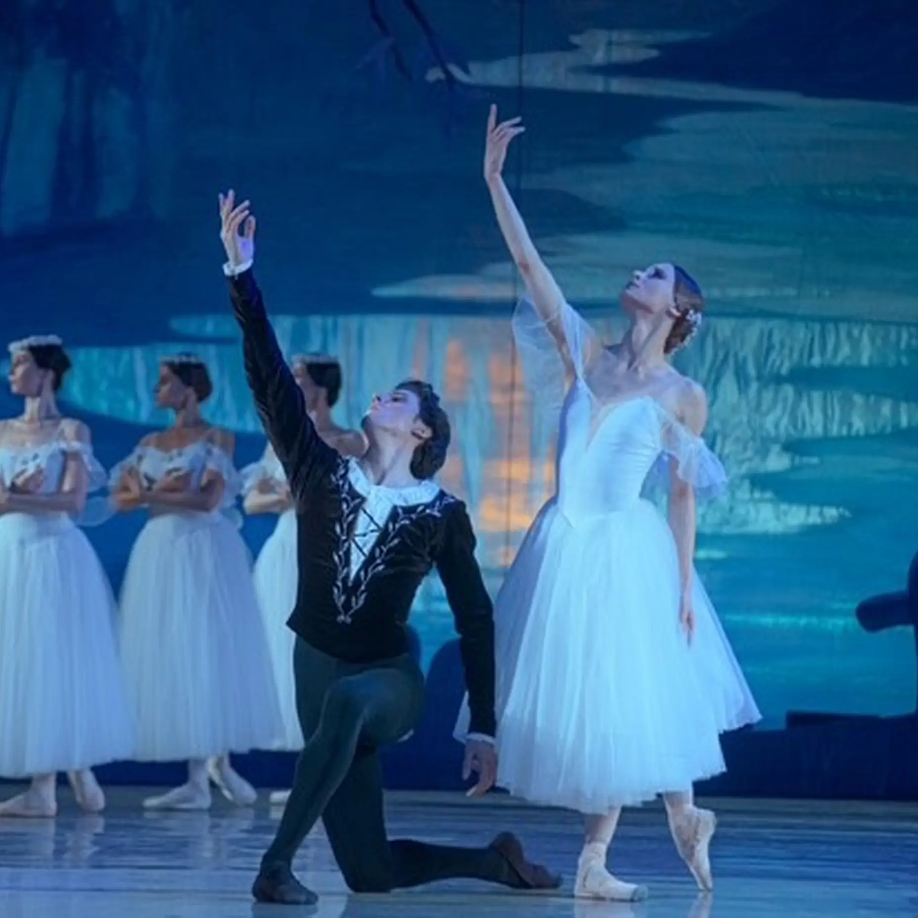Ballettszene auf der Bühne mit Tänzern in weißen Kostümen. Zwei Haupttänzer heben dramatisch die Arme in einer beleuchteten Waldkulisse.
