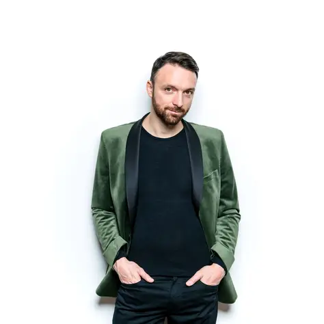 Ein Mann steht vor einem weißen Hintergrund, trägt einen grünen Blazer und ein schwarzes Hemd, Hände in den Taschen.