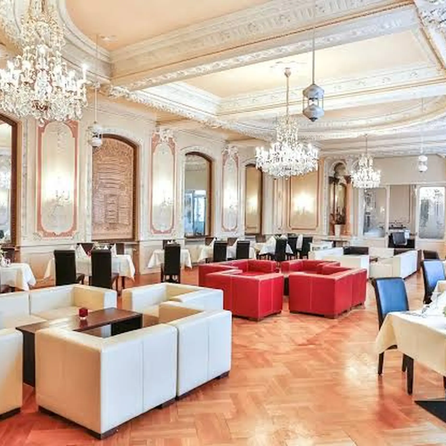 Elegant banquet hall with chandeliers, white sofas, red cushions, and set tables. High ceilings and mirrors on the walls.