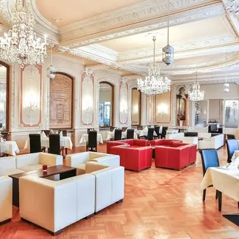 Elegant banquet hall with chandeliers, white sofas, red cushions, and set tables. High ceilings and mirrors on the walls.