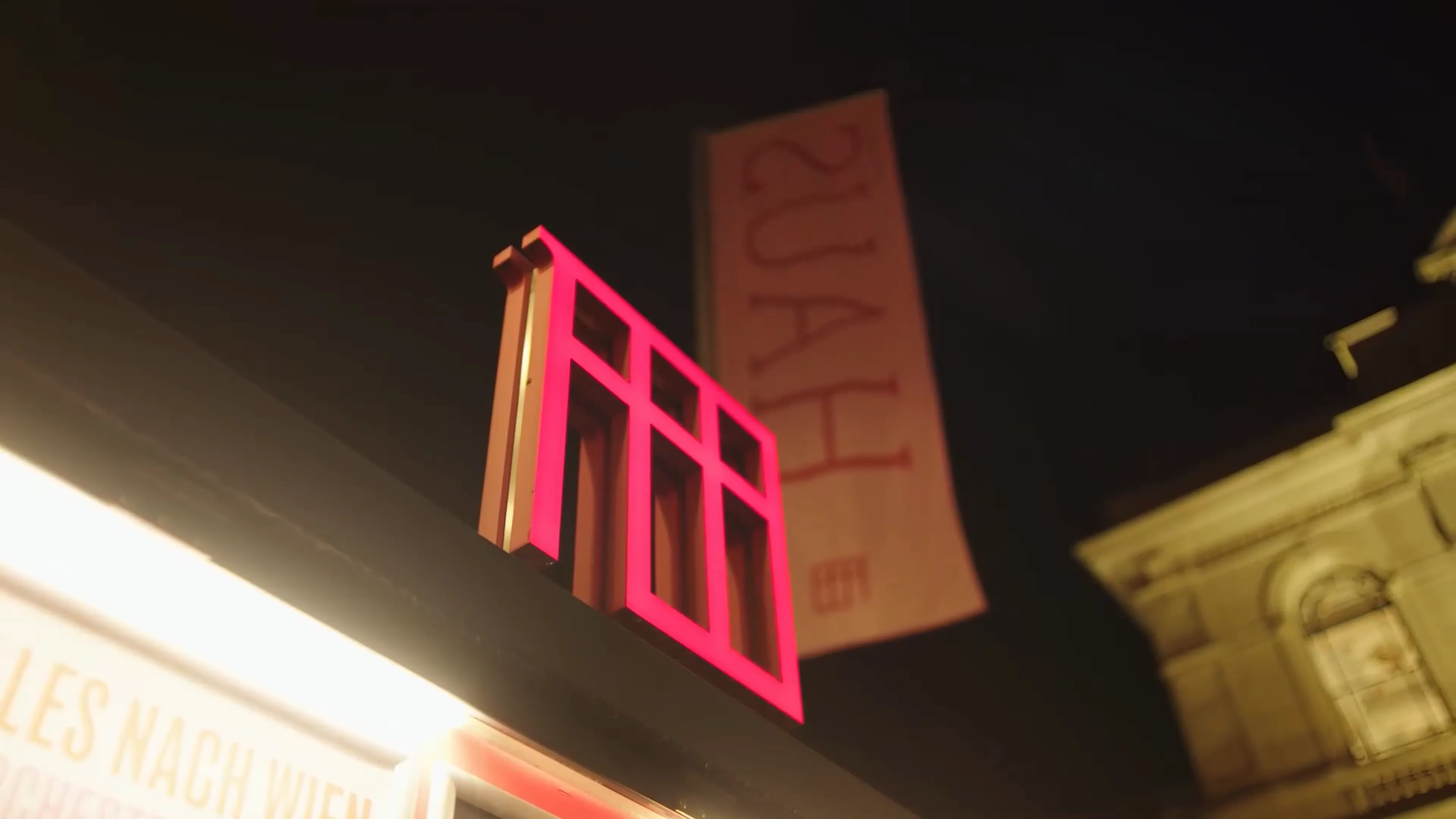 Das leuchtende, rote Logo des Festspielhauses vor dem Gebäude, beleuchtet von unten. Ein vertikales Banner hängt im Hintergrund.
