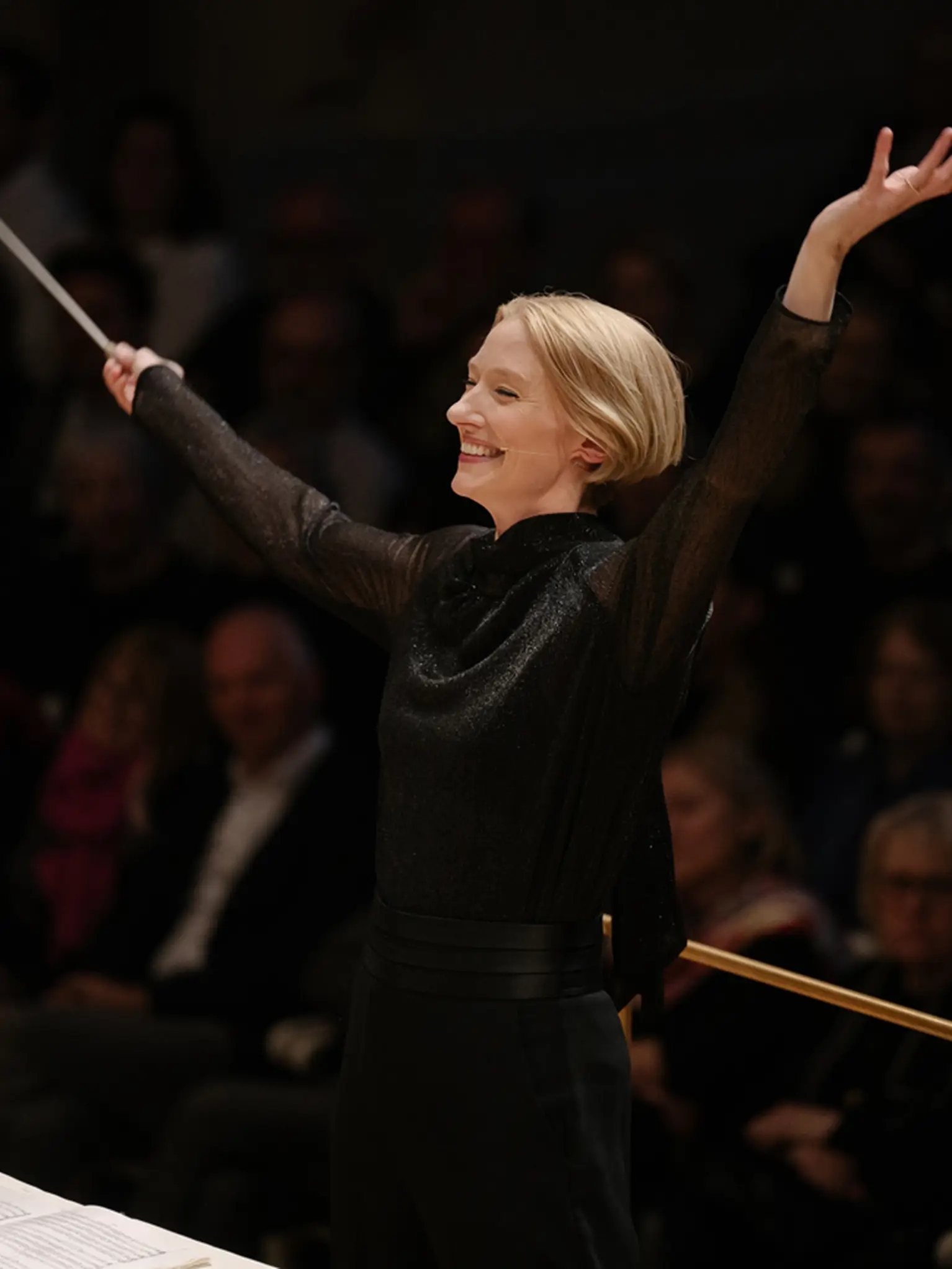 Die Dirigentin Joana Mallwitz in einem schwarzen Kleid dirigiert ein Orchester mit ausdrucksstarken Bewegungen vor einem Publikum.