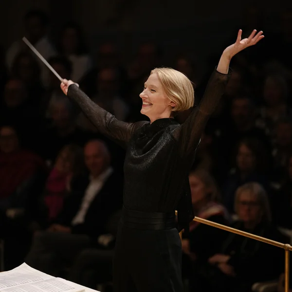 Die Dirigentin Joana Mallwitz in einem schwarzen Kleid dirigiert ein Orchester mit ausdrucksstarken Bewegungen vor einem Publikum.