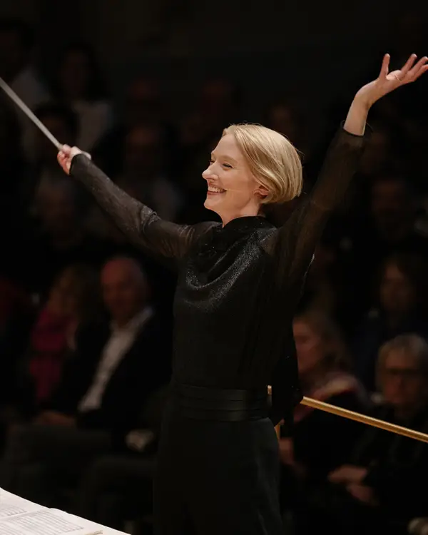 Die Dirigentin Joana Mallwitz in einem schwarzen Kleid dirigiert ein Orchester mit ausdrucksstarken Bewegungen vor einem Publikum.
