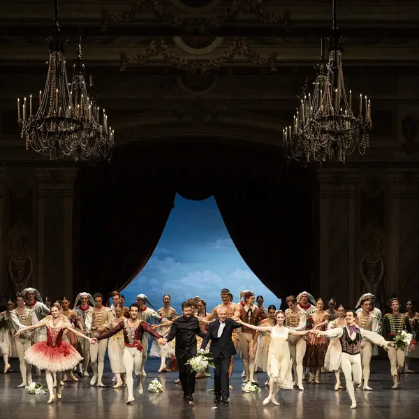 Ballettaufführung auf einer großen Bühne mit vielen Tänzern in farbenfrohen Kostümen, vor einer schönen blauen Kulisse.
