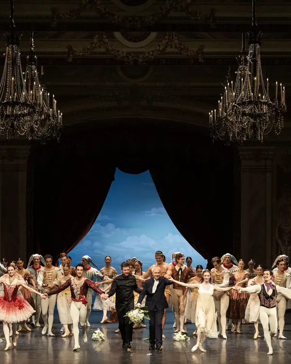 Ballettaufführung auf einer großen Bühne mit vielen Tänzern in farbenfrohen Kostümen, vor einer schönen blauen Kulisse.