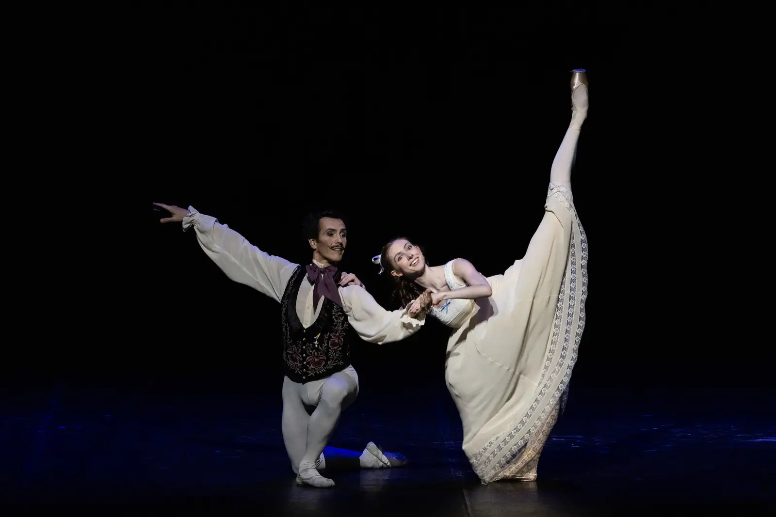 Zwei Balletttänzer auf der Bühne in eleganten Posen. Eine Tänzerin hebt ihr Bein hoch, beide tragen Kostüme.