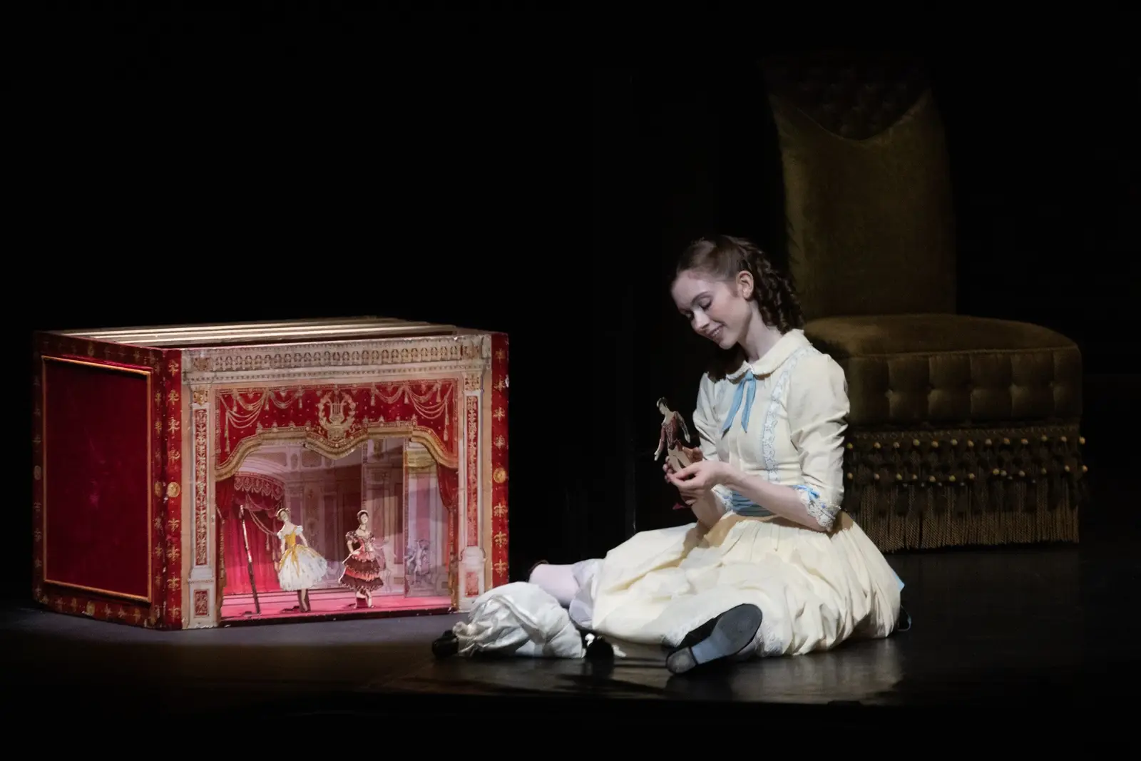 Eine Schauspielerin in einem Bühnenkostüm sitzt auf dem Boden, neben einer kleinen Puppentheaterbühne mit kunstvollen Requisiten.