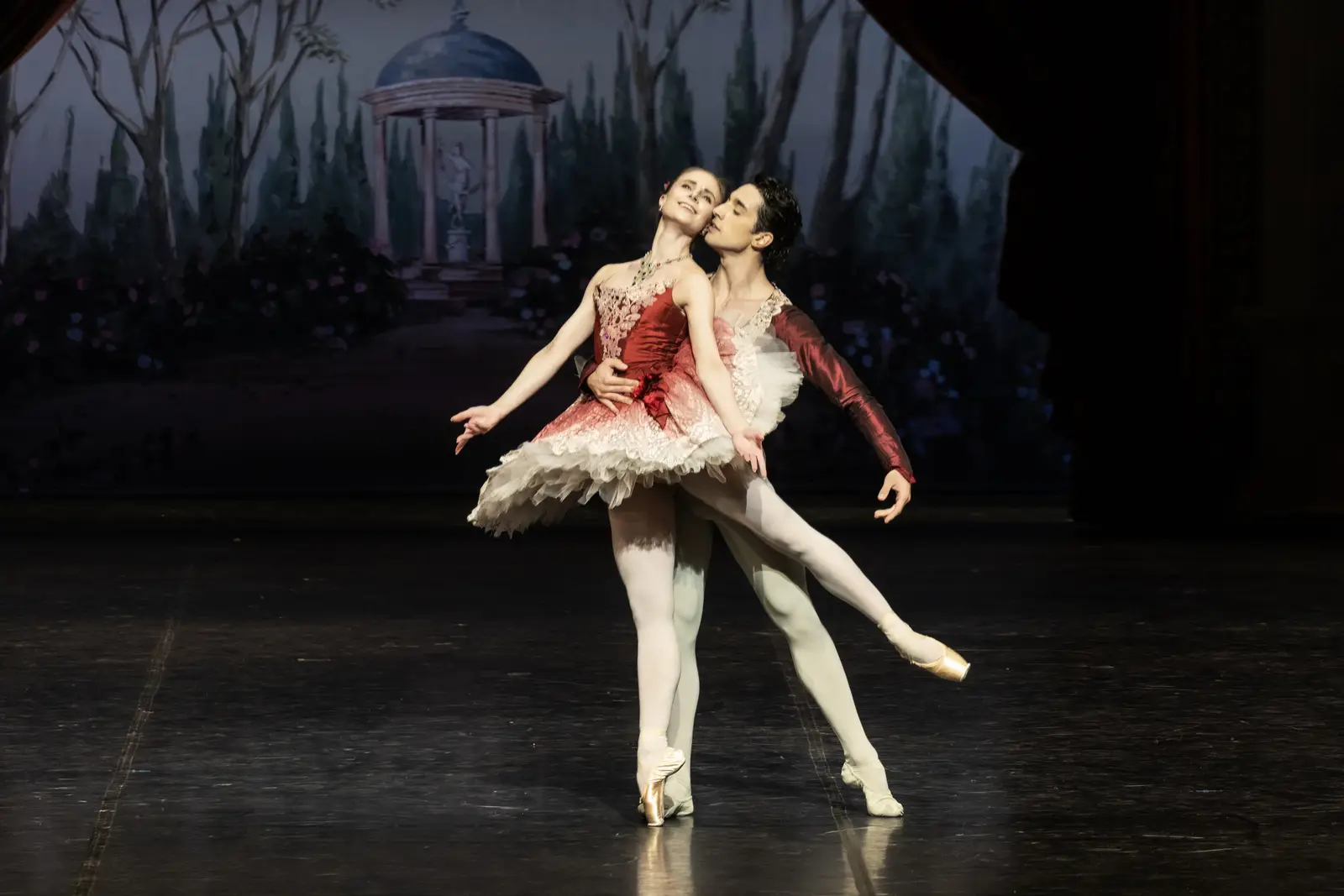 Zwei Balletttänzer führen auf einer Bühne einen klassischen Pas de Deux auf. Sie tragen elegante Kostüme vor einem waldartigen Hintergrund.