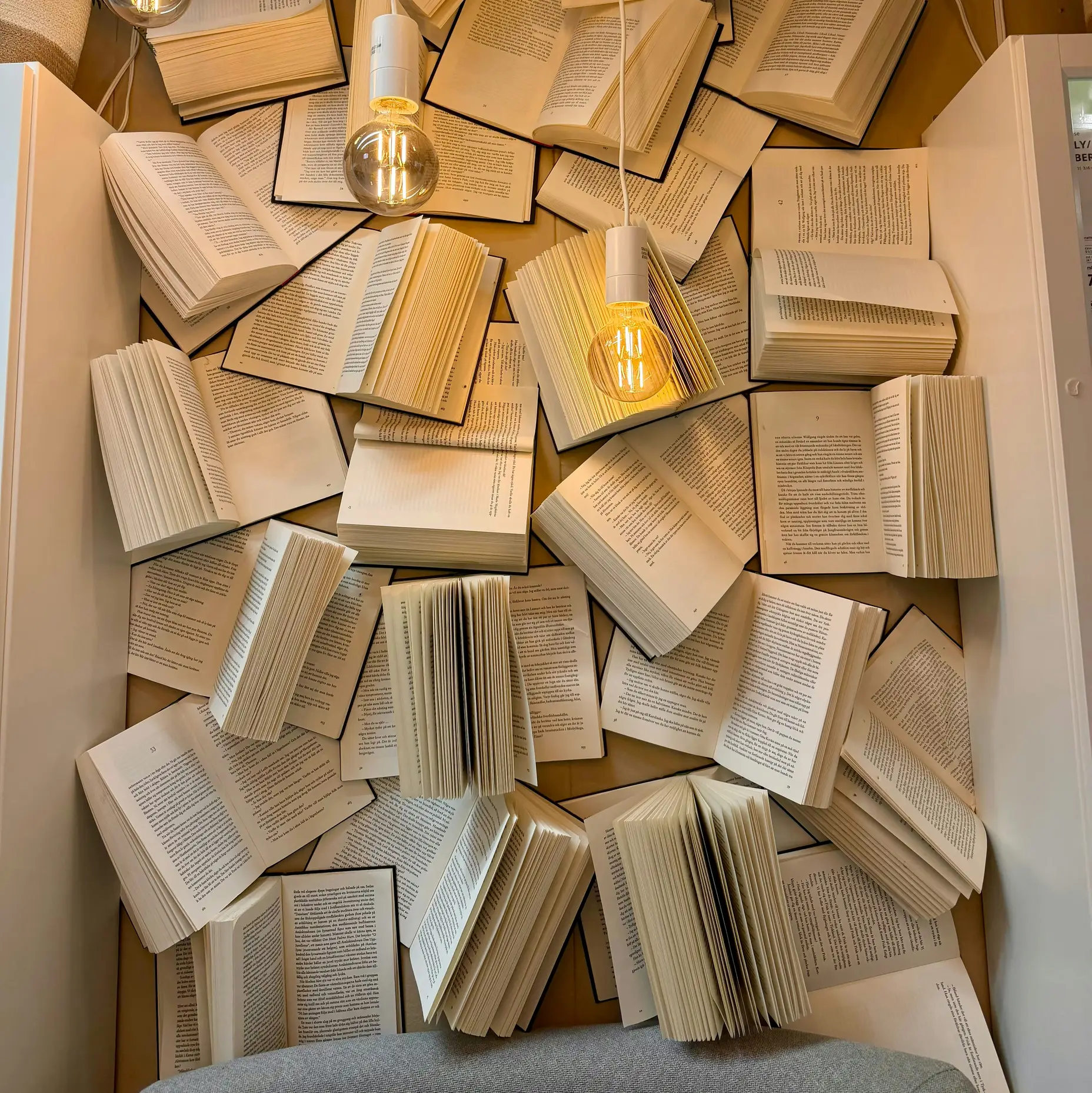 Ein kreatives Wanddisplay zeigt Bücher mit geöffneten Seiten, die aufeinander gestapelt sind, beleuchtet von hängenden Glühbirnen.