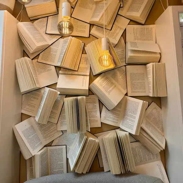 Ein kreatives Wanddisplay zeigt Bücher mit geöffneten Seiten, die aufeinander gestapelt sind, beleuchtet von hängenden Glühbirnen.