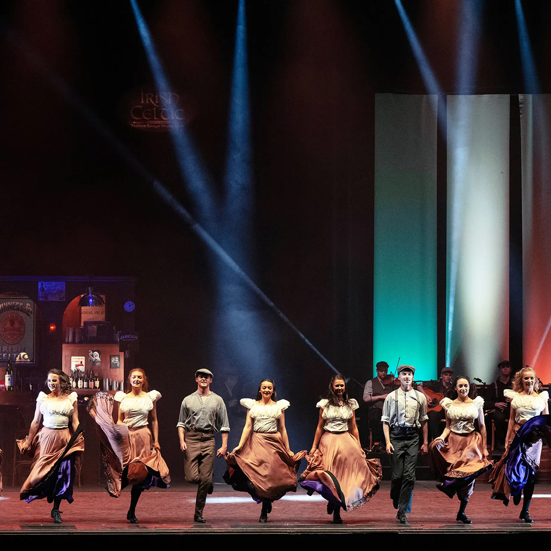 A group of dancers in traditional attire performs energetically on a brightly lit stage, with colorful lights in the background.