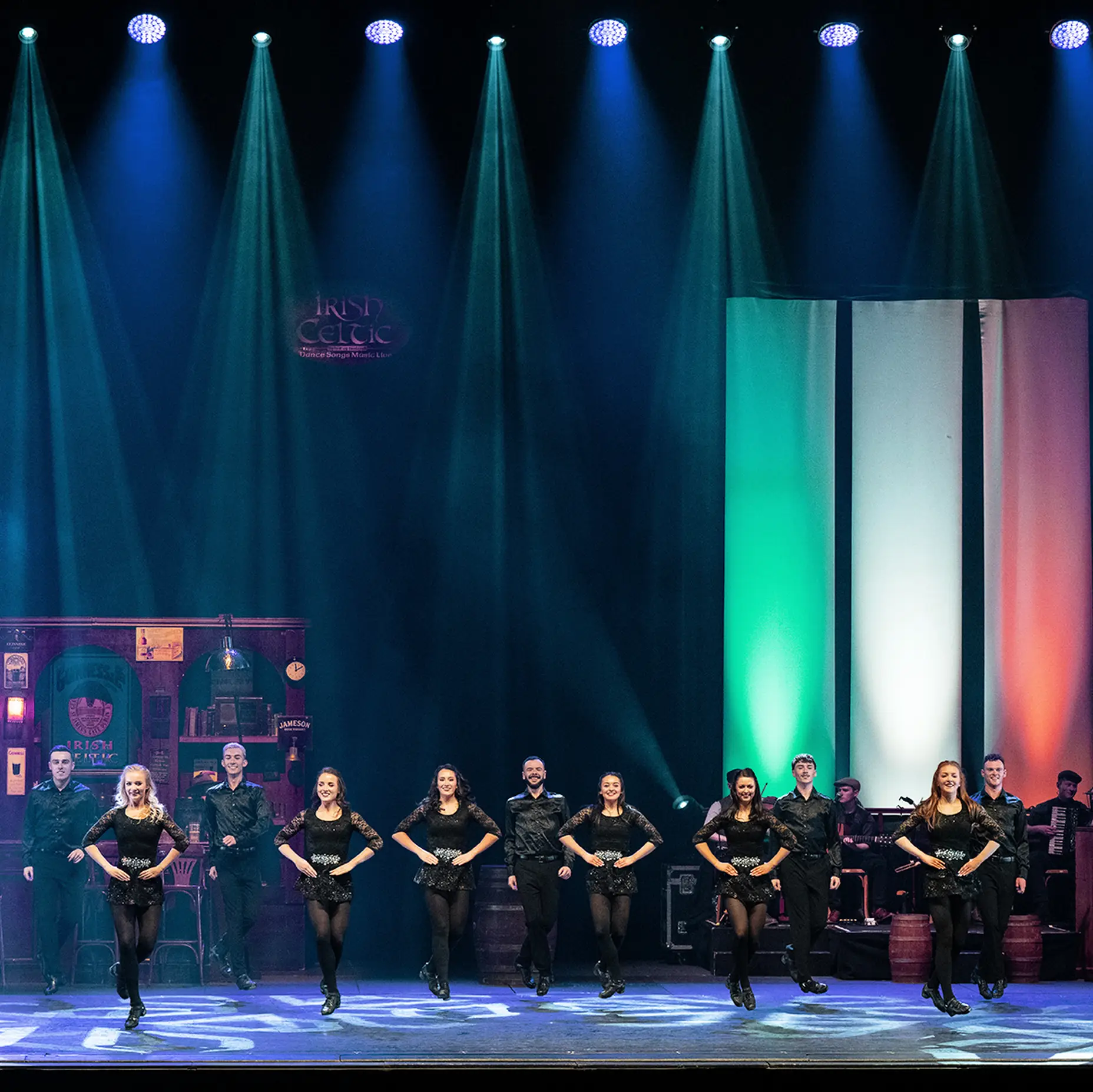 A group of dancers perform Irish step dancing. Behind them, there are bright stage lights and an Irish flag display.