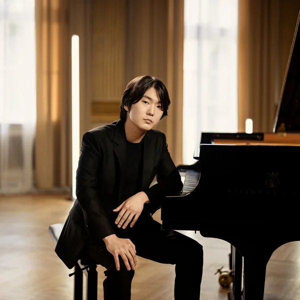 Der Pianist Seong-Jin Cho sitzt entspannt an einem Flügel in einem stilvollen Raum mit weichen Vorhängen und moderner Beleuchtung.