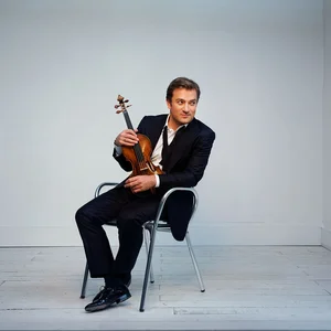 Renaud Capucon sitzt auf einem Stuhl in einem modernen Raum und hält eine Geige. Er trägt einen eleganten Anzug.