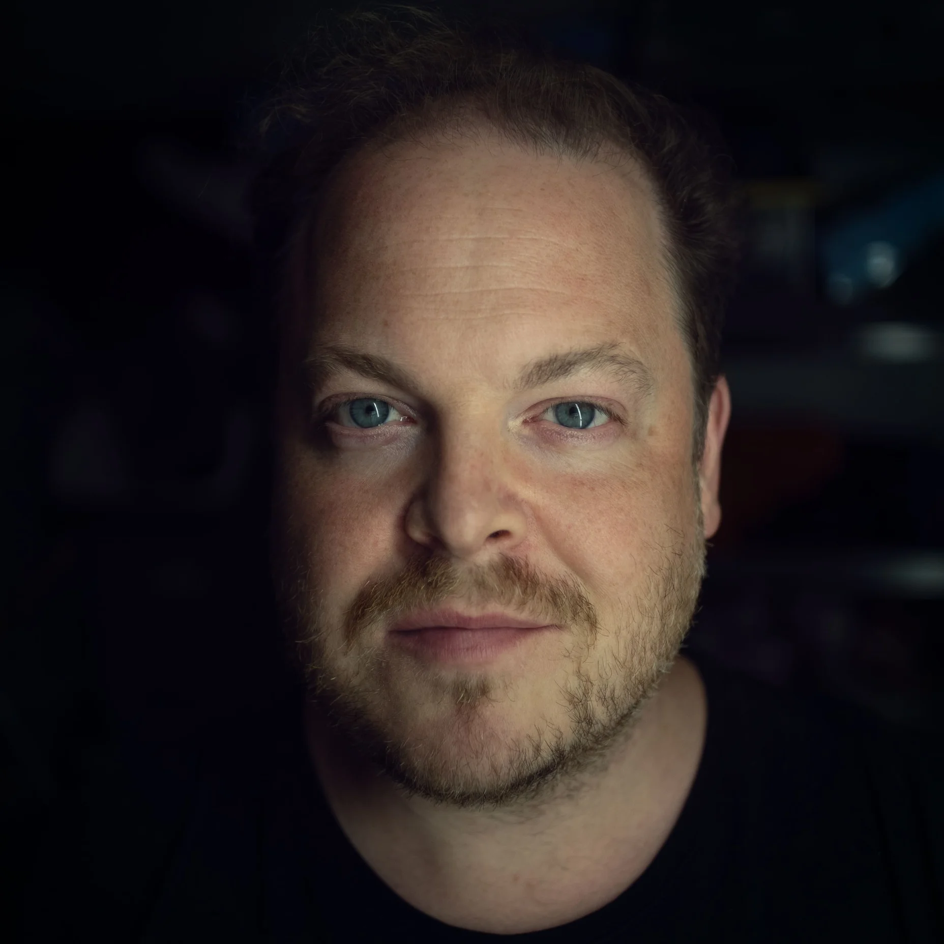 Porträt von Andrew Staples mit bartähnlichem Gesichtsausdruck, beleuchtet in einem dunklen Raum.