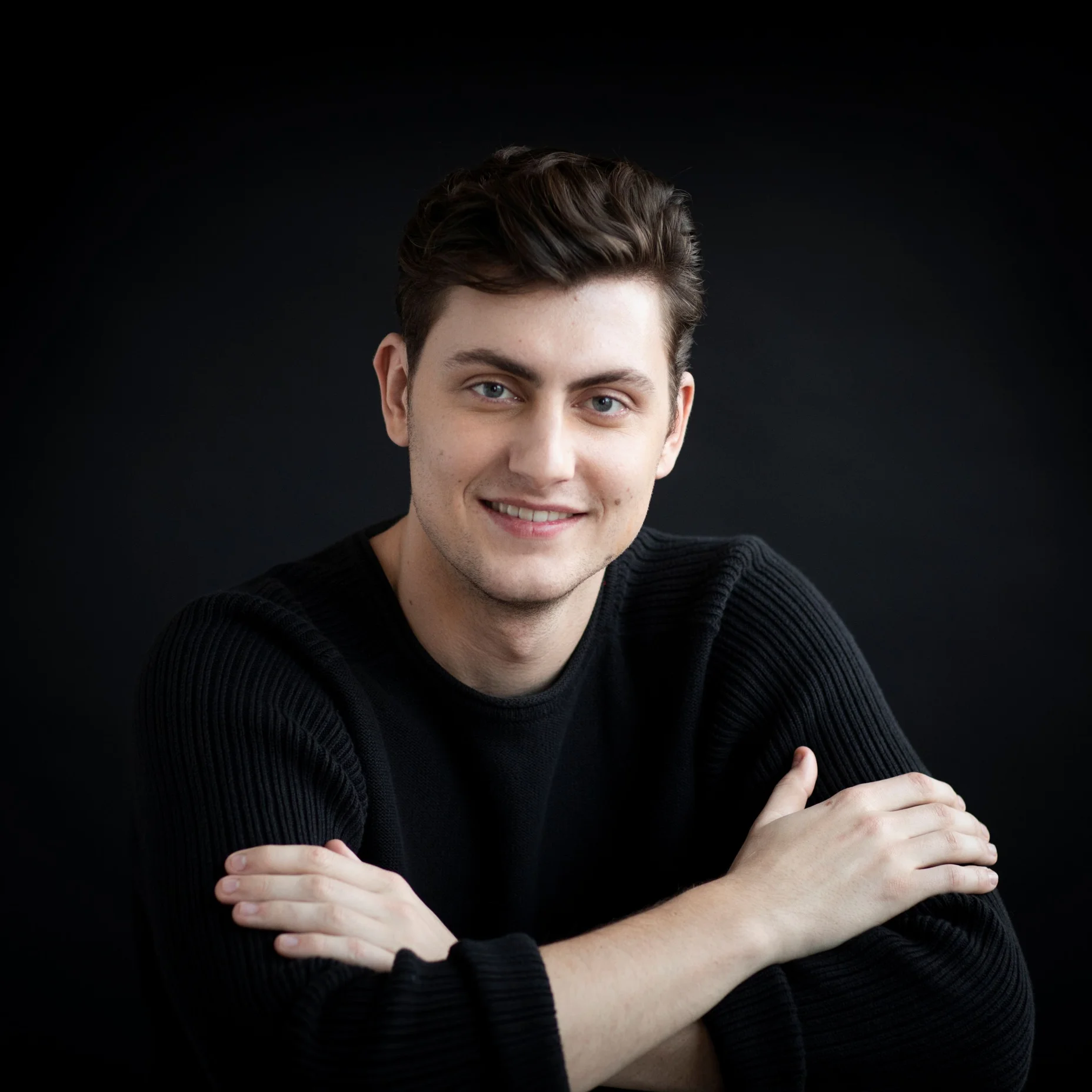 William Thomas in a black sweater smiles with arms crossed in front of his chest. The background is dark.