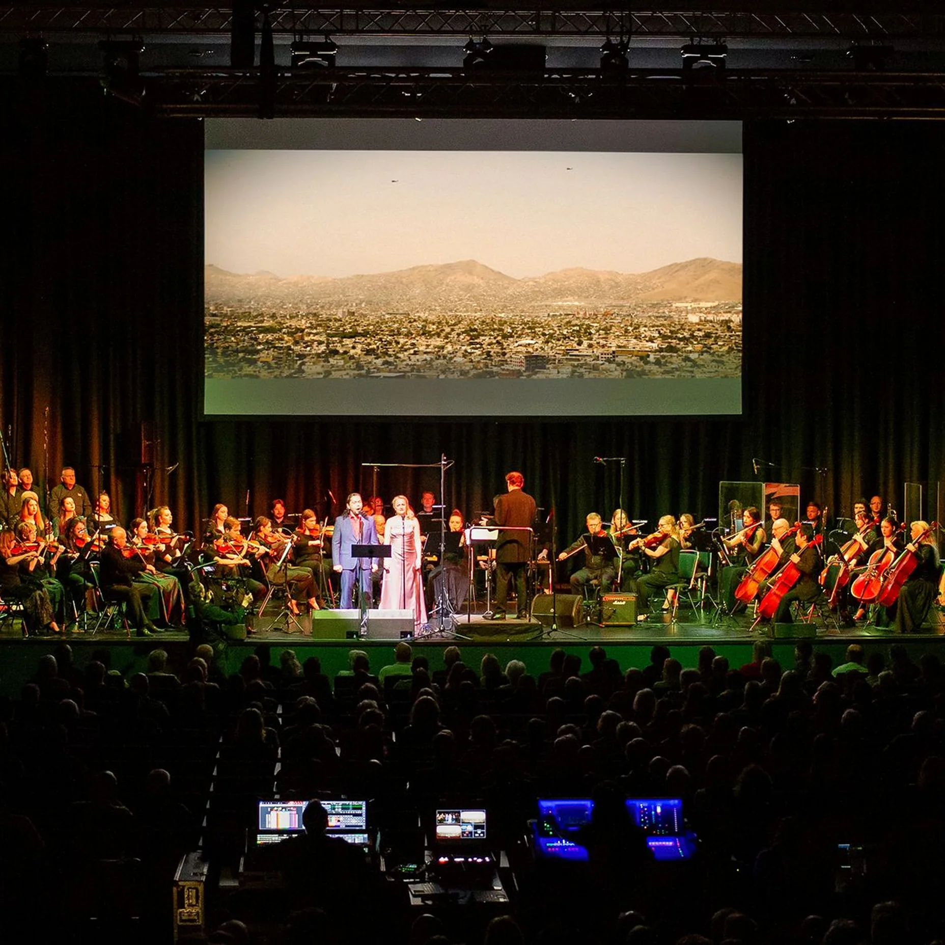 Ein Orchester spielt vor einem großen Bildschirm, der eine Stadtlandschaft zeigt. Sänger stehen in der Mitte der Bühne.