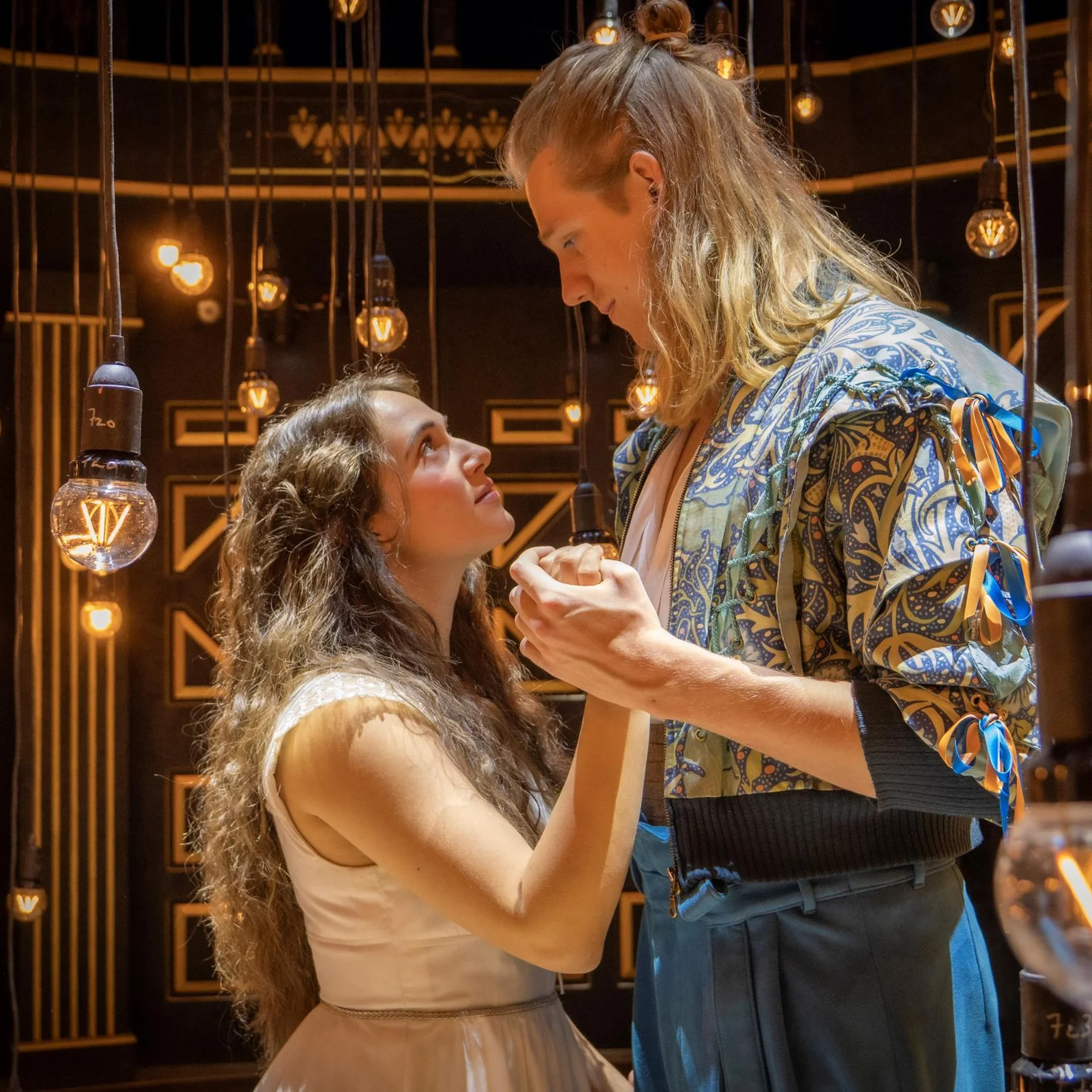 Romeo & Julia in historischen Kostümen tanzen in einem Theater. Um sie herum hängen viele stilvolle Glühbirnen. Die Szene wirkt träumerisch.