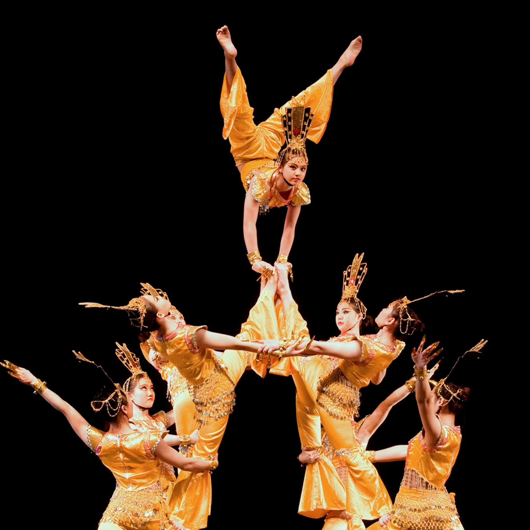 Sechs Künstlerinnen in goldenen Kostümen führen eine akrobatische Tanzpose auf der Bühne auf. Eine Tänzerin balanciert oben.