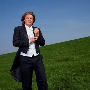 Klaus Florian Vogt en costume marche sur un champ vert sous un ciel bleu clair, ajustant ses boutons.