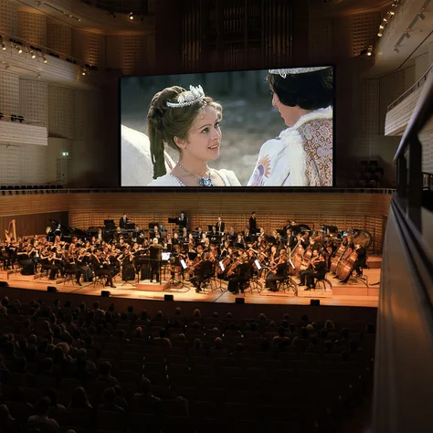 Ein Orchester spielt in einem großen Konzertsaal, während auf einer Leinwand der Film Drei Haselnüsse für Aschenbrödel gezeigt wird. Im Hintergrund sieht man Zuschauer.