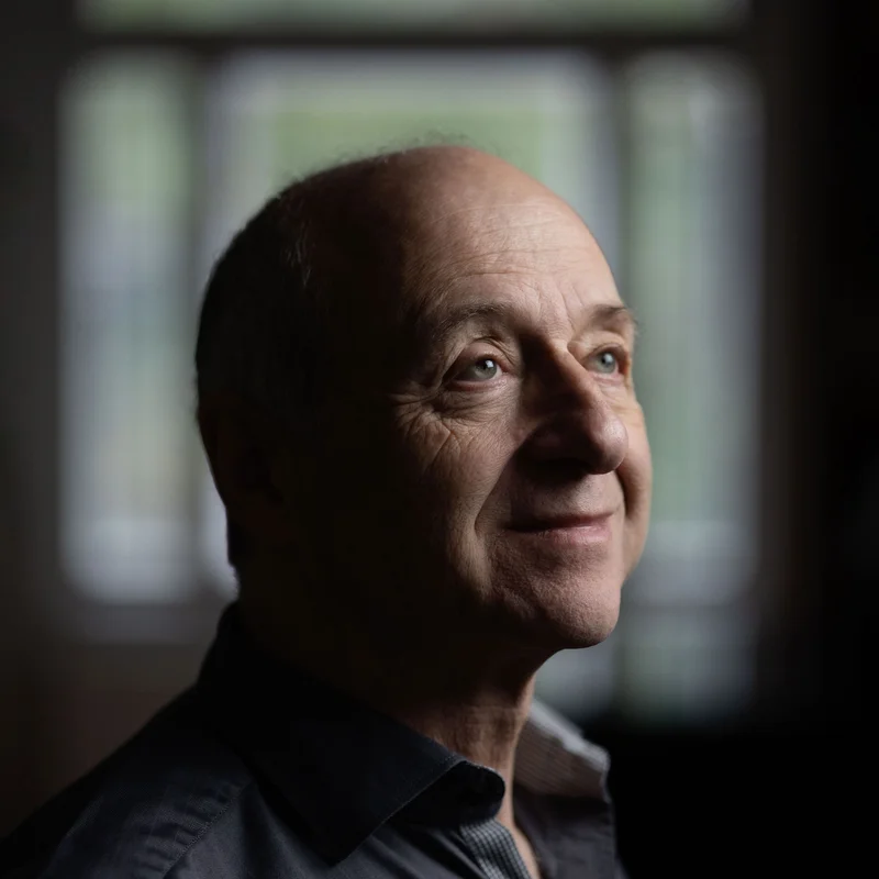 Ivan Fischer gazes thoughtfully upward, standing in front of a window with a blurred background, wearing dark clothing.