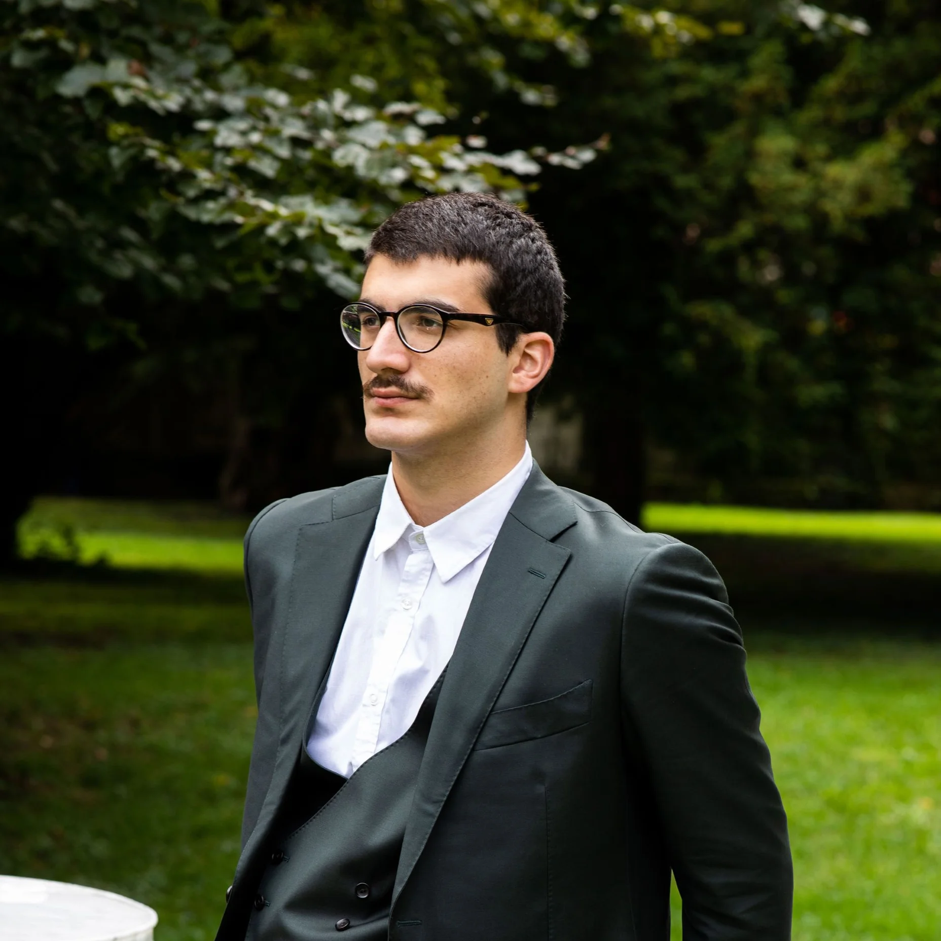 Giorgi Gigashvili in einem eleganten, dunklen Anzug lehnt lässig an einer weißen Steinsäule in einem grünen Garten.